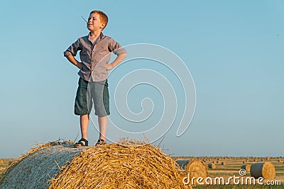 A red-haired boy stands on top of a straw bale on a wheat field Stock Photo