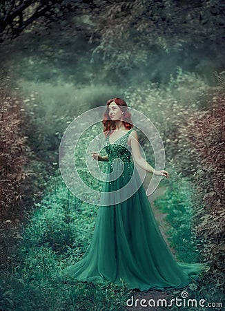 Red-haired girl in a green, emerald, luxurious dress in the floor, with a long train. The princess walks in a fairy Stock Photo