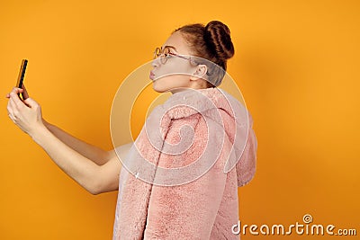A red-haired girl in round glasses stands in profile in a pink coat and takes a selfie, stretching out lips in a kiss Stock Photo