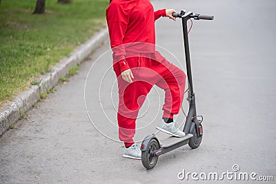 Red-haired girl in a red tracksuit drives an electric scooter. A young woman in oversized clothes rides around the city Stock Photo