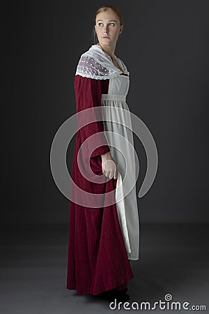 A Regency maid servant wearing a red linen dress with an apron and a lace modesty shawl against a studio backdrop Stock Photo