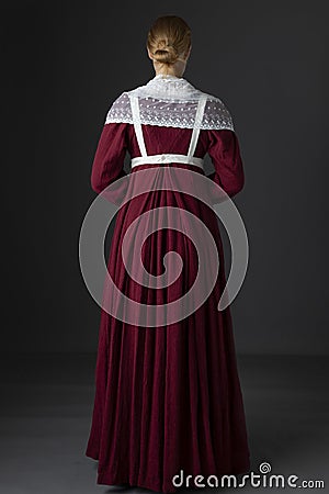 A Regency maid servant wearing a red linen dress with an apron and a lace modesty shawl against a studio backdrop Stock Photo