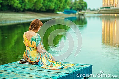 Red-headed woman in bright summer dress with open back sitting on the wooden pier at the river bank Stock Photo