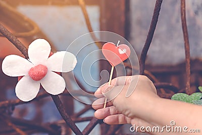 A red heart-shaped candle on woman`s hand and an artificial flower made of white and red candles with bluerd image of rusty steel Stock Photo