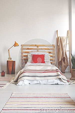 Red pillow on single bed with striped bedding in spacious bedroom interior, real photo with copy space on the empty wall Stock Photo