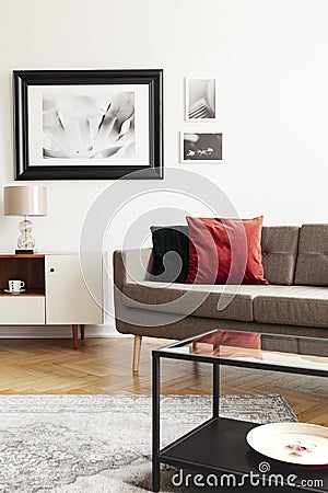 Red pillow on sofa next to white cabinet with lamp in living room interior with posters. Real photo Stock Photo