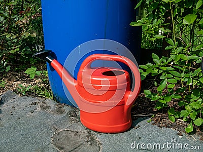 Red, plastic watering on ground can in front of blue, plastic water barrel reused for storing water for watering plants in bright Stock Photo
