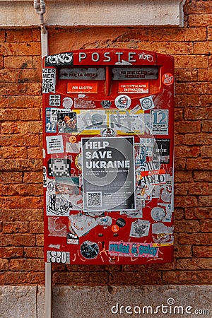 Red postbox adorned with stickers and Help Save Ukraine message flyer against red brick wall Editorial Stock Photo