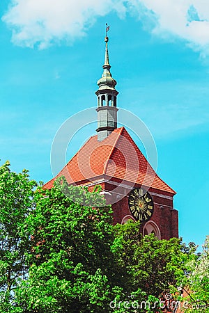 Red-roofed church tower with clock surrounded by green trees against blue sky Stock Photo