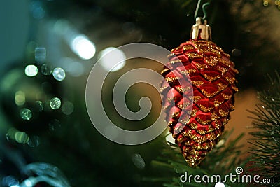 Red shining pine acorn cone hanging on a Christmas tree with blur bokeh background. New Year decoration prop. Stock Photo