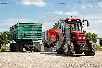 Red tractor with trailer and green trailer alone, parked on a farmyard against blue sky Stock Photo