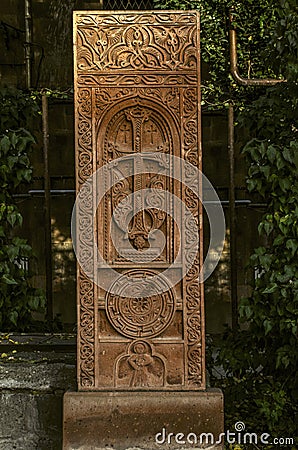 Red tuff khachkar with an arch carved with a cross, a figure of a Saint and a round symbol of eternity on the alley of stone cross Stock Photo