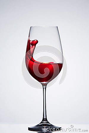 Red wine splashing from the pure elegant wineglass standing against light background with reflection. Stock Photo