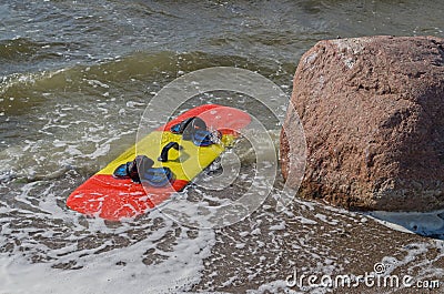 Red and yellow kiteboarding board with carrying handle and slippers attached to it washed up by foaming wave next to large boulder Stock Photo