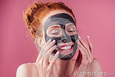 Redhaired ginger teenager girl with black clay mask on her pretty face on pink studio background Stock Photo