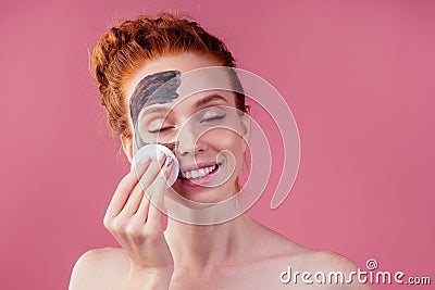 Redhaired ginger teenager girl remove with cotton black clay mask on her pretty face on pink studio background Stock Photo