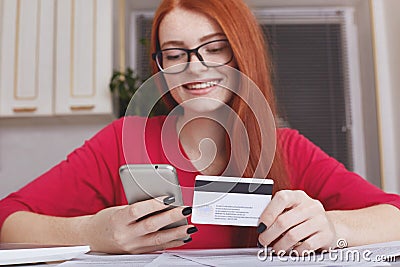 Redhaired pretty female model in eyewear holds smart phone and credit card, makes online purchase or shopping in internet store, h Stock Photo