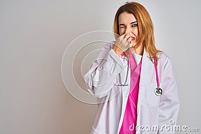 Redhead caucasian doctor woman wearing pink stethoscope over isolated background smelling something stinky and disgusting, Stock Photo