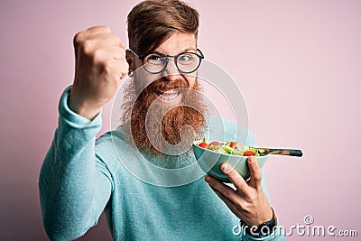 Redhead Irish healthy man with beard eating vegetarian green salad over pink background annoyed and frustrated shouting with Stock Photo