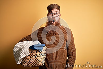 Redhead Irish man with beard doing housework laundry over yellow background scared in shock with a surprise face, afraid and Stock Photo