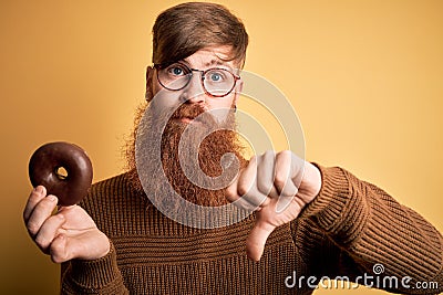 Redhead Irish man with beard eating chocolate donut over yellow background with angry face, negative sign showing dislike with Stock Photo