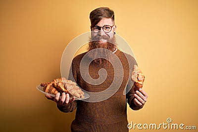 Redhead Irish man with beard eating french croissant pastry over yellow background with a happy face standing and smiling with a Stock Photo