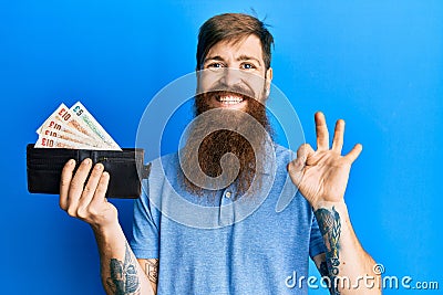 Redhead man with long beard holding wallet united kingdom pounds doing ok sign with fingers, smiling friendly gesturing excellent Editorial Stock Photo