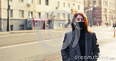 Redhead woman in grey coat and black protective mask standing on an empty sunny street Stock Photo