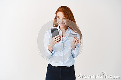 Redhead woman making online order, holding plastic credit card and looking at smartphone screen surprised, white Stock Photo