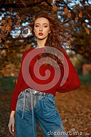 Redhead young woman in a red sweater walks in the park. Autumn beauty portrait of a fashionable Red-haired woman at sunset Stock Photo