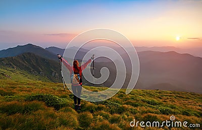 Redheaded girl athlete with a backpack and sticks stands on the green hillocks and looks at high mountain landscapes. Stock Photo