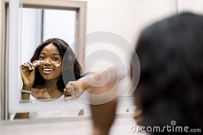 Reflection of pretty smiling African girl in mirror applying cosmetic cream on her face. Female putting moisturizer on Stock Photo