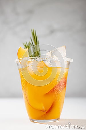 Refreshing juicy peach cocktail with ice, rosemary, sugar rim, fruit slices in misted glass on white wood table, concrete wall. Stock Photo