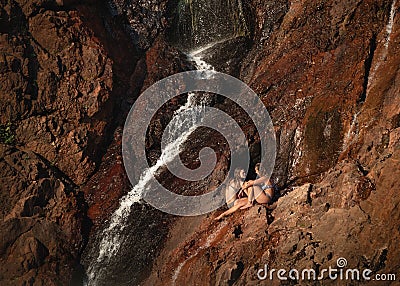 Refreshing summer. Two girls talking next to a waterfall. Sun light at sunset time. Sitting on rock wall. Editing space. Editorial Stock Photo