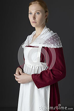 A Regency maid servant wearing a red linen dress with an apron and a lace modesty shawl against a studio backdrop Stock Photo