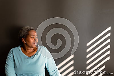 Relaxed african american senior woman sitting at wall in ray of light Stock Photo