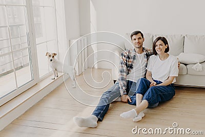 Relaxed couple sit on floor near couch, embrace and smile, dressed in casual clothes and white socks, enjoy domestic atmosphere, Stock Photo