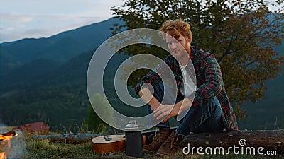 Relaxed tourist camping outside in mountains. Ginger tourist warm hands by fire. Stock Photo