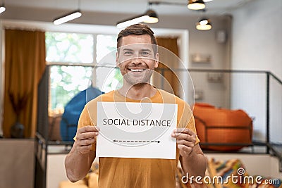 Reopening office safely. Young happy man showing paper with text SOCIAL DISTANCE at camera and smiling, standing in the Stock Photo