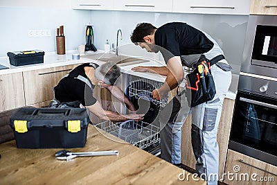 Repair company for you. Aged repairman in uniform fixing dishwasher in the kitchen, while his young colleague helping Stock Photo