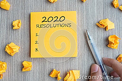 2020 RESOLUTIONS word on yellow note with Businessman holding pen and crumbled paper on wooden table background. New Year start, Stock Photo