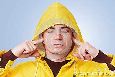 Restful attractive male closes eyes and plugs ears, tries to concentrate and not hear loud noise, wears yellow hood on head, isola Stock Photo