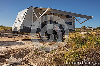 A retired Australian woman sits under the awning of a modern caravan adjacent to a beach in a bright clear day in Australia Stock Photo