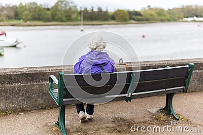 A retired woman sitting alone on a bench sitting out over a river at high tide Editorial Stock Photo