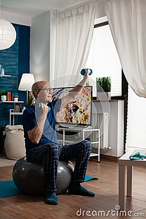 Retirement senior man sitting on aerobics swiss ball raising hand streching arm muscle Stock Photo