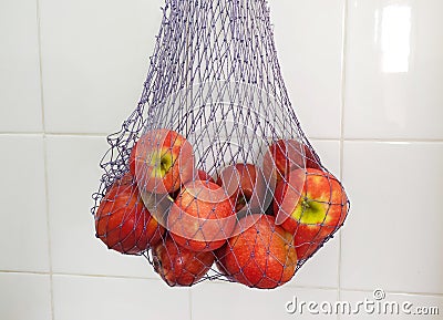 Reusable thread mesh filled with red apples hanging on the ceramic tiles wall. Storage for future use Stock Photo
