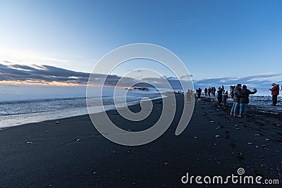 Reynisfjara black sand beach at sunset illuminated by the last rays of the sun from the horizon of the sea with tourists Editorial Stock Photo