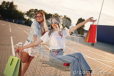 Riding in a shop cart. Two beautiful women in casual clothes are holding bags, outdoors Stock Photo