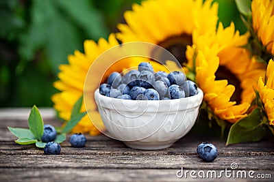 Ripe blueberries in white bowl with sunflower bouquet on wooden table, summer theme Stock Photo