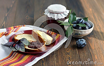 A jar of homemade fruit jam, ripe plums, bread and butter on a red plate. Dark wooden table. Stock Photo